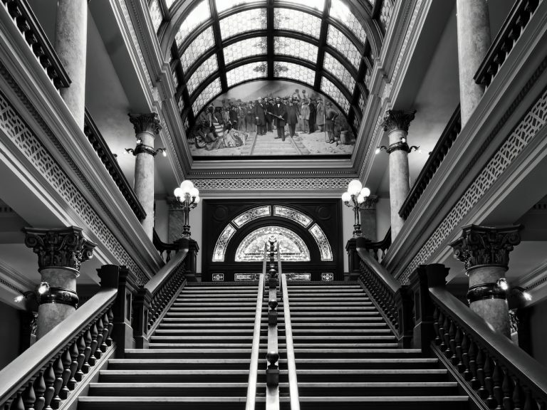 A grand staircase with ornate railings and stained glass windows leads up to a mural depicting a historical event.
